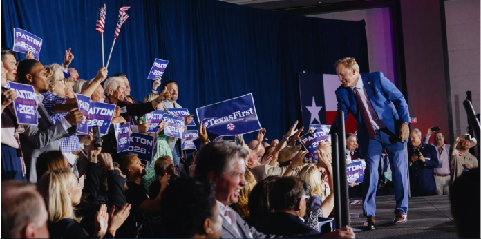 Politician in a blue suit onstage at a rally, reaching toward a cheering crowd holding Paxton 2026 signs and flags nearby.