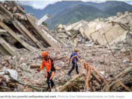 Two rescue workers in orange and blue gear walk amid collapsed buildings and rubble, with a search-and-rescue dog in a mountainous area.
