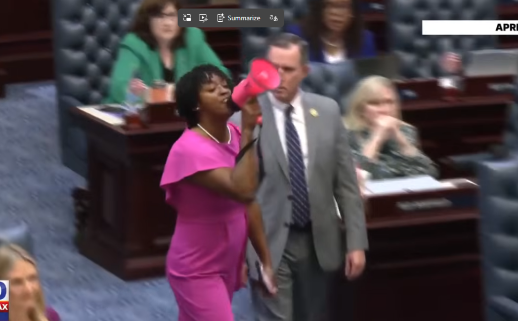 Woman in a Pink Dress Speaks into a Pink Megaphone on a Legislative Chamber Floor, with a Man in a Gray Suit Beside Her. Woman in a Pink Dress Speaks into a Pink Megaphone on a Legislative Chamber Floor, with a Man in a Gray Suit Beside Her.