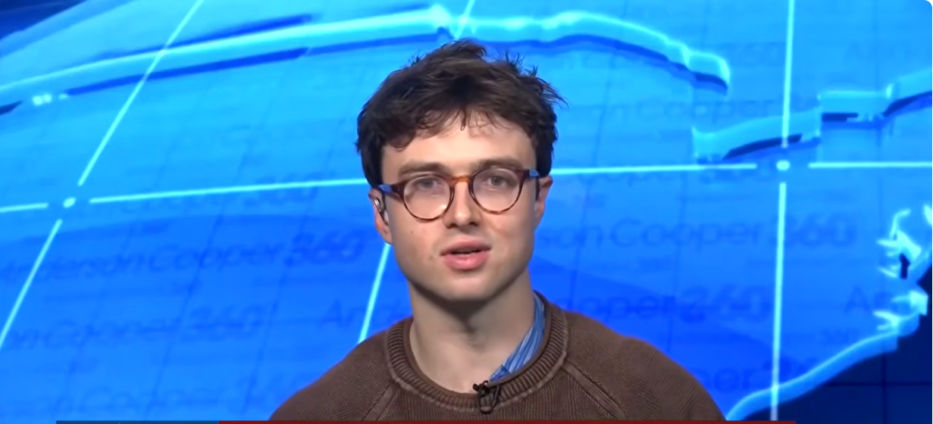 Young Man with Round Glasses Speaking in a Blue Studio Backdrop, Wearing a Brown Sweater with a Mic Clipped to It. Young Man with Round Glasses Speaking in a Blue Studio Backdrop, Wearing a Brown Sweater with a Mic Clipped to It. Young Man with Round Glasses Speaking in a Blue Studio Backdrop, Wearing a Brown Sweater with a Mic Clipped to It. Young Man with Round Glasses Speaking in a Blue Studio Backdrop, Wearing a Brown Sweater with a Mic Clipped to It.