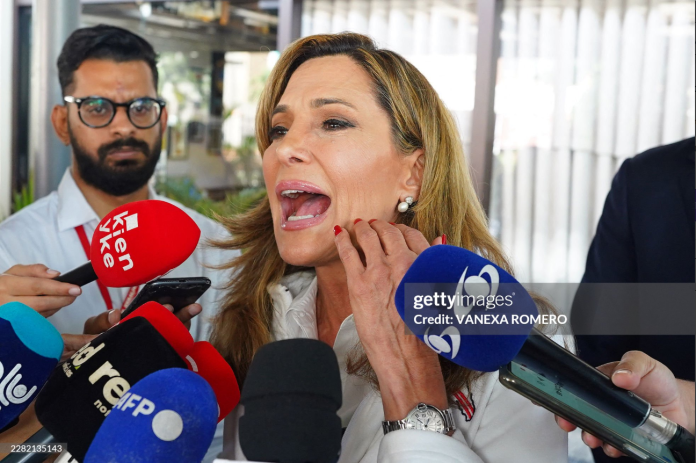 Rep. Elvira Hi Hoe Woman speaking to reporters as microphones from multiple outlets surround her. Behind her, a man with glasses observes.