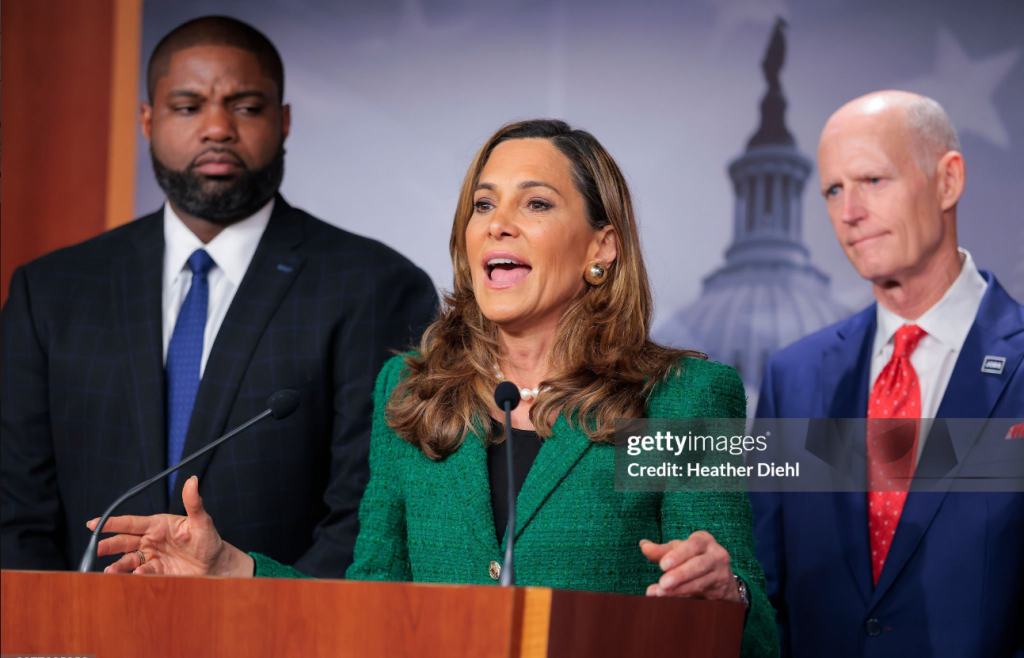 Woman in a Green Blazer Speaks at a Podium, Flanked by Two Suited Men During a Press Conference. Woman in a Green Blazer Speaks at a Podium, Flanked by Two Suited Men During a Press Conference. Woman in a Green Blazer Speaks at a Podium, Flanked by Two Suited Men During a Press Conference. Woman in a Green Blazer Speaks at a Podium, Flanked by Two Suited Men During a Press Conference.