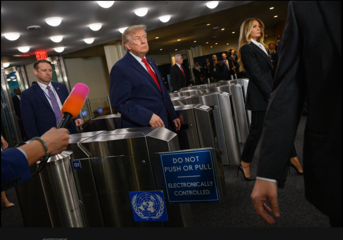 Man in a blue suit walks toward a metal security turnstile as a reporter with a bright microphone stands nearby.